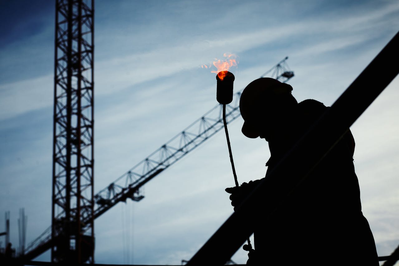 gallery-6 Silhouette of a construction worker using a blowtorch at a building site against a crane-filled skyline.